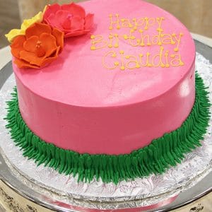 A round pink birthday cake (CB-020C) with "Happy Birthday Claudia" in yellow icing, topped with orange and pink flowers, green icing grass at the base, displayed on a silver platter.
