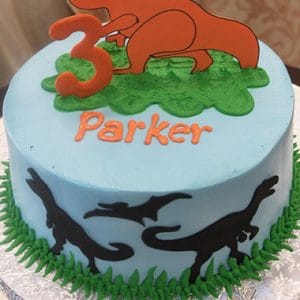 CB-014A is a round blue birthday cake decorated with green grass accents, black dinosaur silhouettes, "Parker" in orange icing, and topped with an orange dinosaur and a number 3.