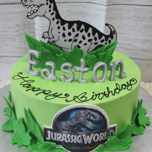 A green CB-014B Jurassic World cake features a cartoon dinosaur topper, green leaves, silver "Easton" letters, and "Happy Birthday" in black icing.
