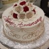 The AN-011 cake is round and white, featuring beige and red roses on top, gold scrollwork on the sides, "Happy Anniversary" in red icing, and is presented on a silver-patterned board.