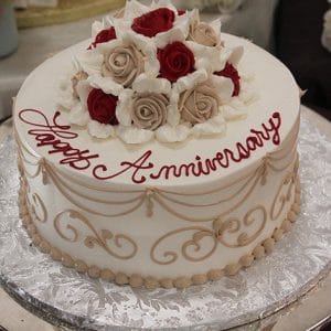 The AN-011 cake is round and white, featuring beige and red roses on top, gold scrollwork on the sides, "Happy Anniversary" in red icing, and is presented on a silver-patterned board.