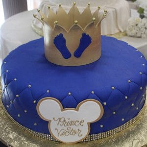 The CB-016E is a round blue cake with gold beads, a gold crown topper, blue baby footprints, and a white and gold Mickey Mouse plaque that reads "Prince Victor.