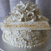 BR-009 is a white birthday cake featuring intricate floral icing, white sugar roses on top, and a white plaque with gold script that reads "Happy Birthday Flora.