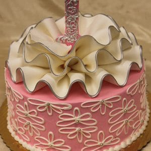 The CB-023B is a pink cake with white icing flowers, ruffled white fondant on top, and a silver number one, presented on a gold cake board.
