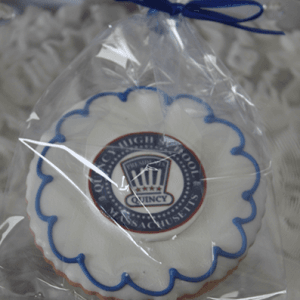 The GC-080C Quincy High School is a round cookie with white icing and a blue scalloped border, wrapped in clear plastic with a blue ribbon. The center features the Quincy High School logo.