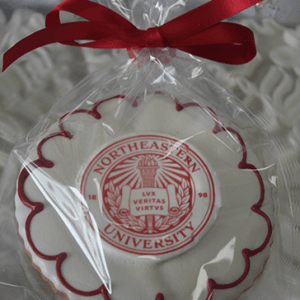 The GC-080G Northeastern University is a round cookie featuring the university's red and white seal, wrapped in clear plastic with a red ribbon, displayed on a white textured surface.