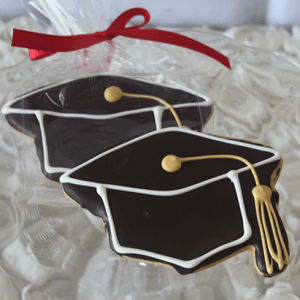The CE-006a graduation includes two graduation cap-shaped cookies decorated with black, white, and yellow icing, wrapped in clear plastic and tied with a red ribbon.