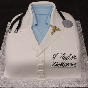 The GC-051 is a cake designed as a doctor’s white coat with a blue shirt, stethoscope, gold caduceus pin, and a J. Taylor Shortsleeve name tag, presented on a patterned silver board.