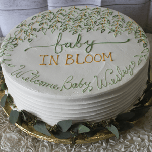 The bb-067 cake features a round, white frosted design with green and yellow floral accents and "baby IN BLOOM Welcome Baby Wesley" written in matching icing. It sits on a gold board with greenery at the base.