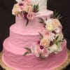 The WD-018 is a three-tiered cake with pink frosting on the bottom two layers, white frosting on top, adorned with pink, white, and peach roses plus greenery, and presented on a gold board.