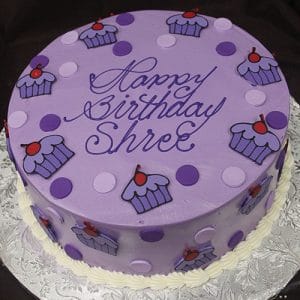 The CB-025E is a round purple birthday cake decorated with polka dots and cupcake designs, featuring "Happy Birthday Shree" in cursive icing on top, presented on a silver patterned board.