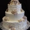 WD-030 is a three-tiered white wedding cake with textured icing, topped with realistic edible seashells and starfish, displayed on a silver-patterned base against a dark background.