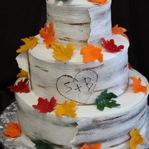 WD-023 is a three-tiered cake styled as birch bark with colorful autumn leaves, a heart with S + B carved on the middle tier, and is presented on a silver foil-covered base.
