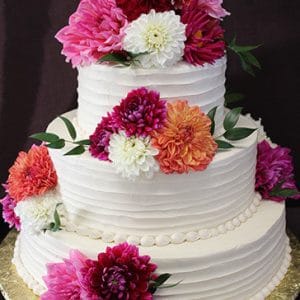 The WD-021 is a three-tiered white cake with piped icing, topped with vibrant dahlias in red, pink, orange, and white, presented on a gold cake board against a dark background.