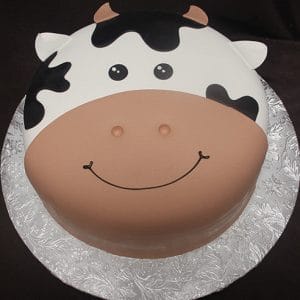 The CB-012C is a round cake designed as a cute cow's face, featuring black and white patches, small ears, nostrils, and a smiling mouth. It is presented on a silver floral-patterned cake board.
