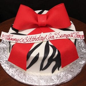 The BD-037B Zebra Bow Red cake features black-and-white zebra stripes, a large red fondant bow, and a white banner with red "Happy Birthday Ba Duong & Trang" script, displayed on a silver-patterned board.