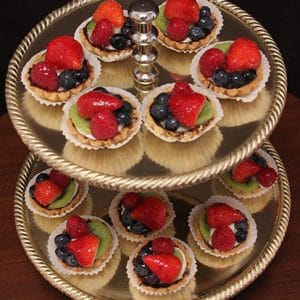 A two-tiered gold tray holds Fruit Tarts, each in a white paper cup and topped with strawberries, blueberries, raspberries, and kiwi slices. The tray rests on a dark wooden surface.