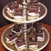 A two-tiered gold tray displays Chocolate Cake Squares, each topped with chocolate frosting and white icing designs, arranged in white paper cups.