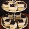 A two-tiered silver tray showcases Boston Cream Diamonds—layered pastry squares with creamy white filling, glossy dark chocolate glaze, and a decorative white swirl on each piece.