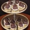 A two-tiered silver stand displays Chocolate Chapeaux truffles, each topped with a chocolate decoration and set in a white paper cup, resting on a dark brown surface against a black background.