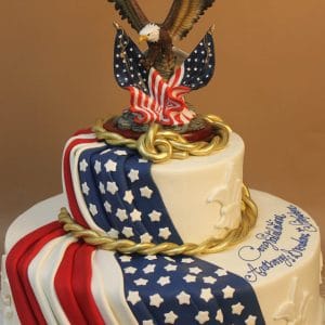 The TC-152 is a two-tiered cake featuring an American flag design, gold rope accents, white stars, and a bald eagle figurine with spread wings on top. A congratulatory message decorates the side.