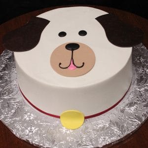 CB-012D is a round cake designed as a dog's face, featuring chocolate ears, black eyes, a brown nose, a smiling mouth, and a yellow collar on a silver-patterned cake board.