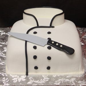 GC-047 is a cake designed to resemble a chef’s white jacket with black buttons and trim, finished with a lifelike edible knife laid diagonally across the front.