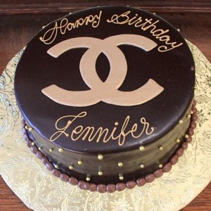 The BD-027A Designer Gold Ganache is a round chocolate cake with a glossy finish, topped with a gold Chanel logo and "Happy Birthday Jennifer" in gold writing, and decorated with gold pearls around the edge.