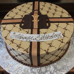 The BD-033D Designer pocket book cake features tan and brown icing, a fondant handbag, and a Happy Birthday message on top, designed to look like a luxury gift box atop a textured silver cake board.