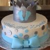 BB-027 is a round white cake decorated with blue and silver polka dots, a large blue bow, a silver crown topper featuring blue baby feet, and "Baby Boy Lizandro" written in blue.