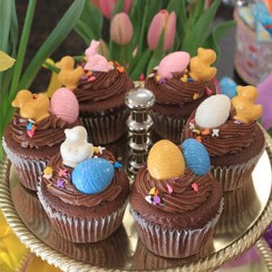 EA-040 Display Easter Decor chocolate cupcake with white filling features colorful Easter-themed toppers like candy eggs, ducks, and bunnies, arranged on a gold cake stand with pink tulips and flowers in the background.