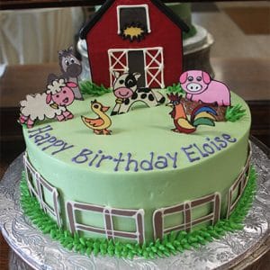 CB-012K: A round birthday cake with green icing, red barn, farm animal toppers (sheep, horse, cow, pig, chick, rooster), and "Happy Birthday Eloise" in purple.