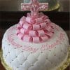 A round white cake (RC-077) with a quilted pattern, pearl-like accents, pink fondant ribbons, a decorative cross, and "Christening Day Eleanor Jane" written in pink.