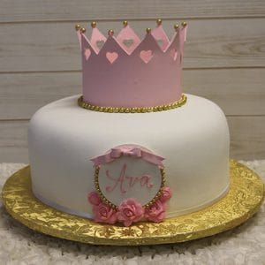 BB-030 is a white round cake with a pink crown topper, gold bead trim, and pink roses. It sits on a gold board with a plaque featuring the name Ava in pink, set against a light wood background.