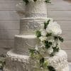 The WD-032 is a four-tiered white wedding cake featuring intricate piping, pearl-like accents, rhinestone bands, and cascading white flowers with greenery, displayed against a light wooden background.