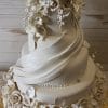 The WD-033 is a three-tiered white wedding cake adorned with realistic white fondant flowers, leaves, and draped icing, presented on a gold cake board with a light wooden background.