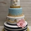 The WD-059 is a four-tier cake featuring navy blue and white striped fondant, nautical rope details, beach-themed seashell and grass accents, pink fondant flowers, all set on a gold base against a wood backdrop.