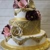 The WD-029 is a three-tier cake featuring gold ornate patterns, gold ribbon accents, and large edible flowers in burgundy, pink, and cream. It rests on a gold textured base against a light wood background.