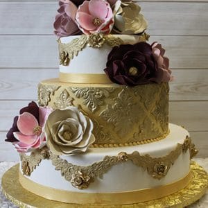The WD-029 is a three-tier cake featuring gold ornate patterns, gold ribbon accents, and large edible flowers in burgundy, pink, and cream. It rests on a gold textured base against a light wood background.