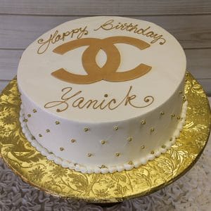 The BD-027D Designer White/Gold cake is a round white birthday cake featuring gold bead accents, a large gold Chanel logo on top, "Happy Birthday Zanick" in gold icing, and is displayed on a gold-patterned cake board.
