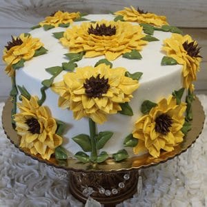 The THX-032 is a round cake with smooth white icing, decorated with large yellow sunflowers and green leaves. It sits on a gold stand with hanging crystals, set against a light wooden background.