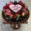 VC-010 is a chocolate cake topped with strawberries, pink heart decorations, and a heart-shaped plaque that says "Happy Valentines Day," presented on a decorative cake stand.