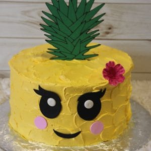 CB-020A is a round cake designed as a cute pineapple, featuring yellow frosting, a fondant green leaf topper, big cartoon eyes, pink cheeks, a smiling mouth, and a pink flower accent on the side.
