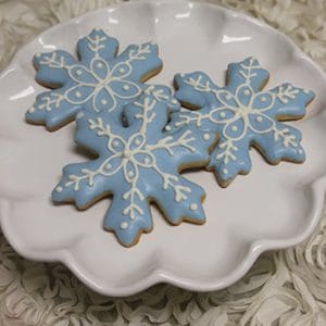 Three AP-075 snowflake cookies with light blue and white icing are arranged on a white, scalloped-edge plate placed on a textured, cream-colored surface.