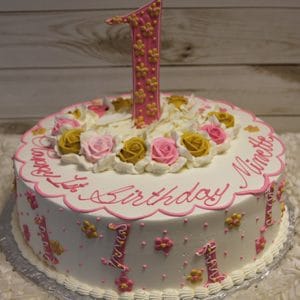 CB-023C: Round white cake with pink and gold flowers, pink and gold number ones, topped with a large pink number one and roses. “Happy 1st Birthday Nimette” is written in pink icing.