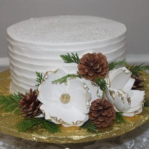 The BD-131 features a white frosted, ridged cake adorned with white and gold-edged flowers, pinecones, and green foliage, presented on a gold-colored board.