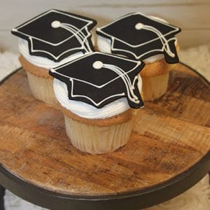Three GC-073 Chocolate cupcakes with white filling, topped with black graduation cap cookies, are displayed on a round wooden cake stand.