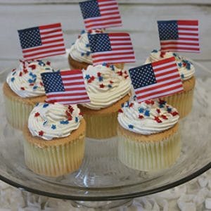 Six CC-037 chocolate cupcakes with white chocolate mousse filling on a glass plate, topped with white frosting, red, white, and blue star sprinkles, and small American flag toothpick toppers. Background is light and rustic.