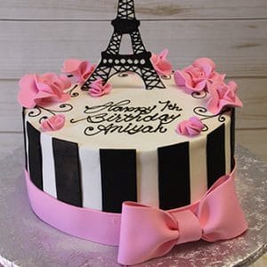 The CB-015A is a round cake with black and white vertical stripes, pink fondant flowers, a large pink bow, a black Eiffel Tower topper, and “Happy 1st Birthday Aniyah” written in script.