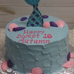 The CB-021D is a blue birthday cake decorated with pink and purple seashells, a mermaid tail topper, and "Happy Sweet 16 Autumn" written in pink icing. The cake sits on a silver board.